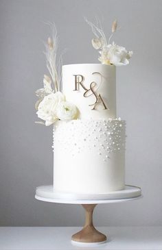 a white wedding cake with flowers and initials on top, sitting on a pedestal in front of a gray background