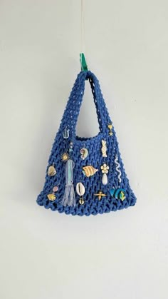 a blue crocheted purse hanging from a hook on a white wall with beads and seashells attached to it