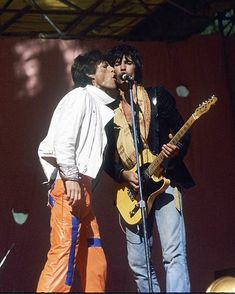 two men standing next to each other while one man sings into a microphone and the other plays an electric guitar