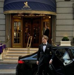 a man in a tuxedo walking past a black car