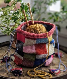 a multicolored knitted bag with yarn and knitting needles in it sitting on a table next to a potted plant