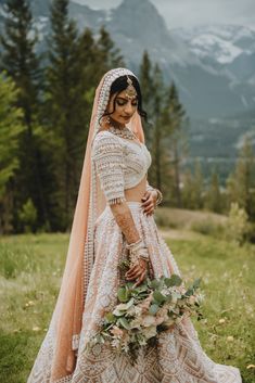 a woman in a white and peach wedding dress standing on grass with mountains in the background