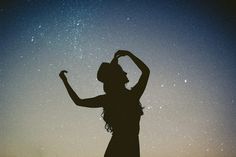 the silhouette of a woman with her arms up in the air, against a night sky filled with stars