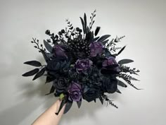 a hand holding a bouquet of purple flowers and greenery in front of a white wall