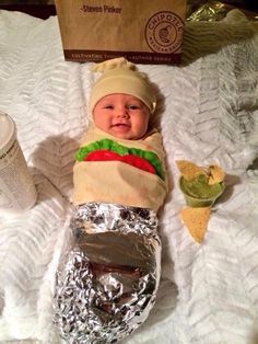 a baby is dressed up like a hot dog and wearing a taco hat while sitting on a bed