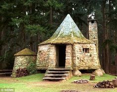 an old stone house in the woods with people sitting on the grass near it and trees