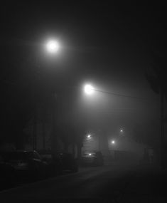 cars are parked on the street at night with lights shining in the foggy sky