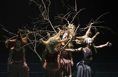 four women in long dresses are performing on stage with bare branches behind them and one woman is holding her arms out to the side