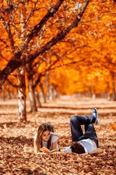 two people laying on the ground in front of trees with leaves all over them and one person reading a book