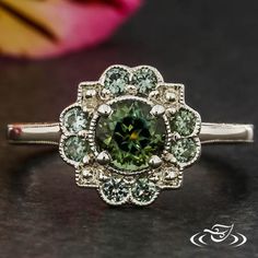 a fancy ring with green and white stones on it's side, sitting next to a pink flower