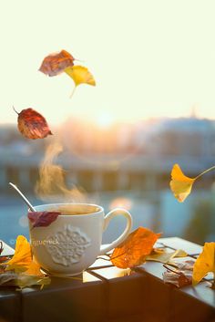 a cup of coffee on a table with autumn leaves falling from the sky in the background