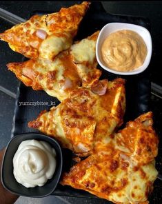 several pieces of pizza on a black tray with dip and sauce in small bowls next to it