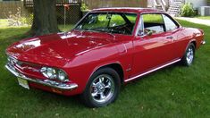 an old red car parked in the grass near a tree and fenced yard area