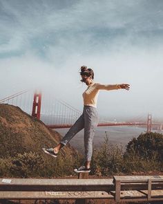 a woman standing on top of a wooden bench next to a body of water with a bridge in the background