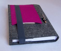 a gray felt case with a pink cover and black strap is holding a small notebook