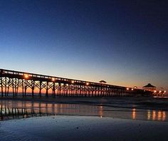 a pier lit up at night with lights reflecting in the water and people walking on the beach