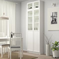 a white dining room with chairs and a bookcase next to a table in front of a window