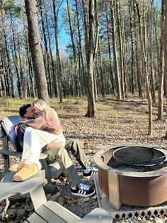 a man and woman sitting next to an open fire pit in the woods with their feet on each other