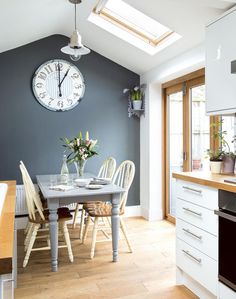 an image of a kitchen setting with blue walls and white furniture in the background, there is a large clock on the wall above the table