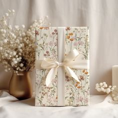 a gift box wrapped in white paper with a bow and flowers on the side next to it