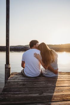 a man and woman are sitting on a dock looking out at the water as the sun sets