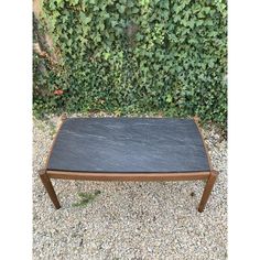 a black slate top coffee table with wooden legs on gravel ground next to green bushes