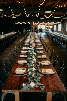 a long wooden table with place settings and greenery on the top is surrounded by lights