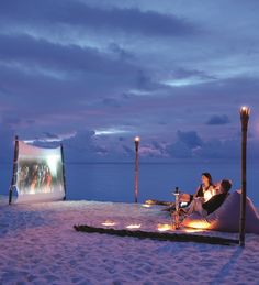 two people are sitting on the beach watching movies and drinking wine at night with candles in front of them