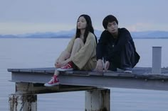 two people sitting on a dock next to the water at dusk, one person wearing red converse shoes