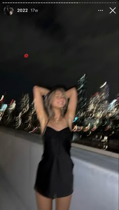 a woman standing on top of a roof next to a tall building at night with her hands behind her head