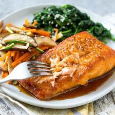 a white plate topped with salmon, vegetables and gravy next to a fork