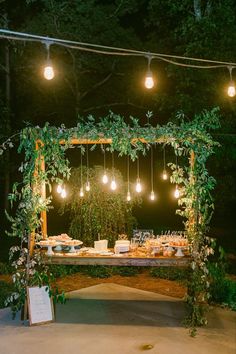 an outdoor wedding reception setup with greenery and hanging lightbulbs at night