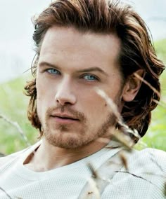 a man with long hair and blue eyes looks at the camera while standing in tall grass