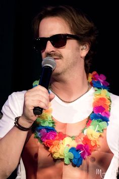 a shirtless man holding a microphone in his right hand and wearing flower leis around his neck