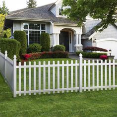 a white picket fence in front of a house with flowers and bushes on the lawn