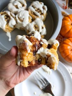 a person holding up a piece of cinnamon roll with icing on it next to some pumpkins