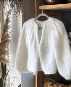 a white knitted sweater hanging on a wooden shelf next to a cup and plant