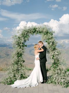 a bride and groom kissing in front of an arch made of greenery with mountains in the background