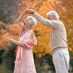 an older man and woman dancing in front of trees with autumn leaves on the ground