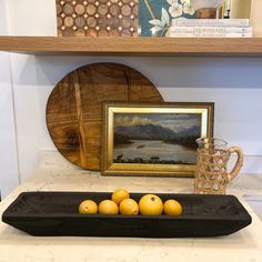 there are some oranges in a black tray on the counter next to a painting