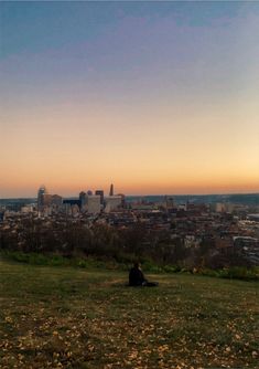 a man sitting on top of a grass covered field next to a tall city skyline