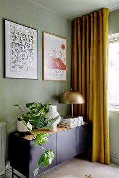 a living room with green walls and yellow drapes on the windowsill, a black sideboard topped by a potted plant next to a gold lamp