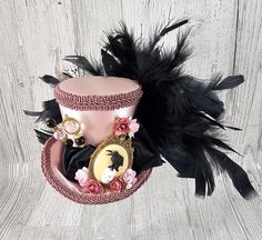 This dramatically gorgeous little hat would be a great addition to any outfit to add a bit of whimsy and fun. It is made from a beautiful dusty pink fabric, trimmed in dusky pink conso gimp braid and fully lined. It features a black satin ribbon band with sculptural ribbon accents, a raven on a skull cameo in a gold setting, pink paper flowers, lovely hat pins, and black feathers that move wonderfully.  The last image is to show the size of the hat only.  Size: This is one of our medium sized mini top hats. The wee top hat itself measures about 3 ¾ inches tall, and the brim is 4 ½ inches wide. The embellishments may extend further off the hat. Check out the last picture to see the size on a person. To see other hats this size, check out this link: https://www.etsy.com/shop/TheWeeHatter?sea Raven On Skull, Dusty Pink Fabric, Top Hat Fascinator, Pink Paper Flowers, Skull Cameo, Mini Top Hats, Alice In Wonderland Mad Hatter, Wonderland Mad Hatter, Little Hat