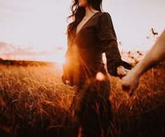 a woman holding the hand of a man who is walking through tall grass at sunset