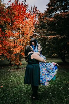 two women hugging each other in front of trees with red leaves on them and one woman wearing a blue dress