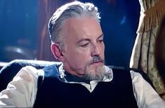 an older man with grey hair and beard wearing a black vest sitting in a chair