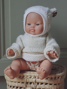 a baby doll sitting in a basket wearing a white knitted sweater and matching diaper