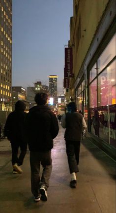 two men walking down the sidewalk in front of a building at night time with city lights behind them