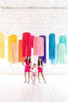 three girls standing in front of colorful streamers hanging from the ceiling and holding hands behind their backs