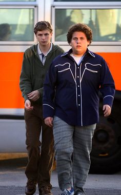 two young men walking down the street in front of a large orange and white bus